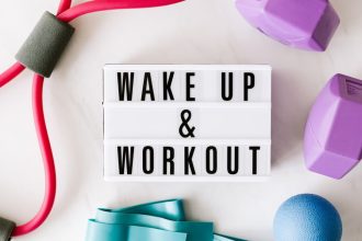 Top view closeup composition of wake up and workout inscription on light box surface among expanders and dumbbells and massage ball on white background
